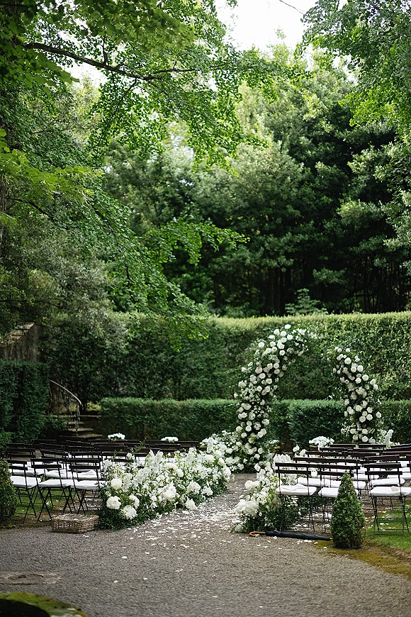Ceremony setup for an outdoor wedding ceremony with a white floral arch, petal-strewn gravel aisle, and black chairs in a hedge garden