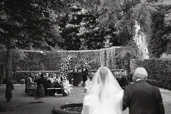 Wedding processional with bride walking down aisle in a long veil toward groom in tux beneath a floral arch in a garden courtyard