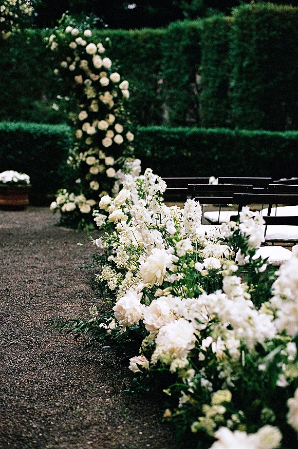 Ceremony aisle decor with wedding aisle flowers and white blooms lining a gravel path, flanked by black chairs and garden hedges