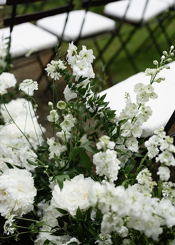 Ceremony aisle florals with wedding aisle flowers in white blooms and greenery lining a grass lawn beside black folding chairs