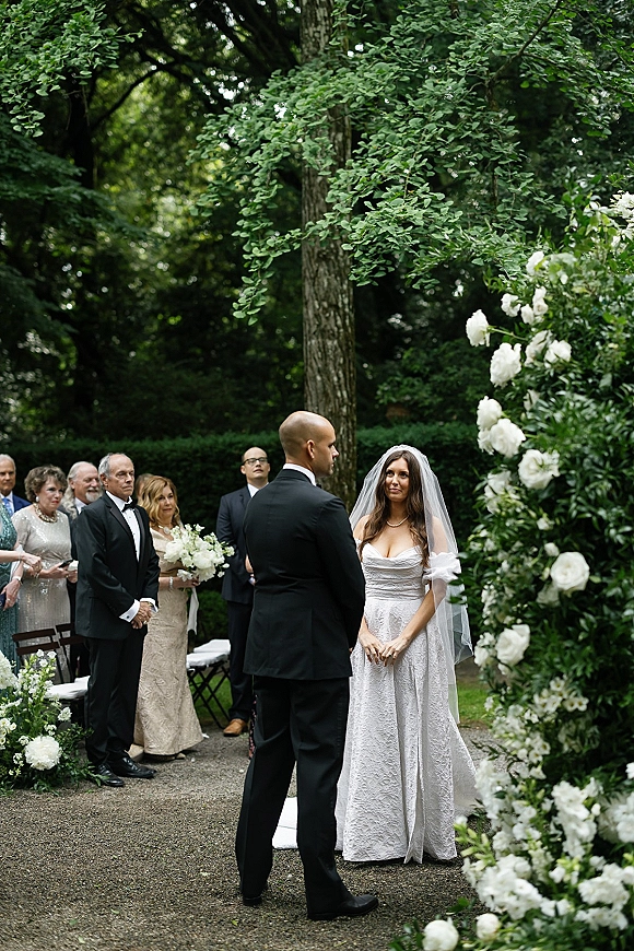 Wedding vows during an outdoor garden ceremony, bride in veil and lace dress with groom in tux beneath a white floral arch on gravel aisle