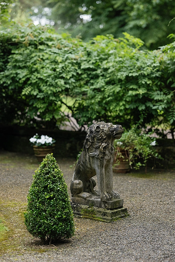 Garden statue featuring a stone lion statue on a pedestal beside a topiary shrub, with potted white flowers on a gravel path amid trees