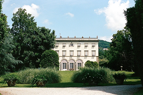 Wedding venue exterior of a white villa with arched doors, shutters, and potted plants, set by manicured lawn and distant hills