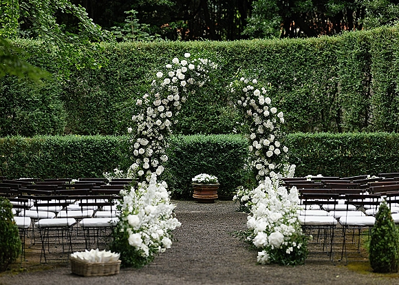 Ceremony setup with an outdoor wedding ceremony floral arch of white roses and greenery, aisle flowers, and chairs on a gravel garden path