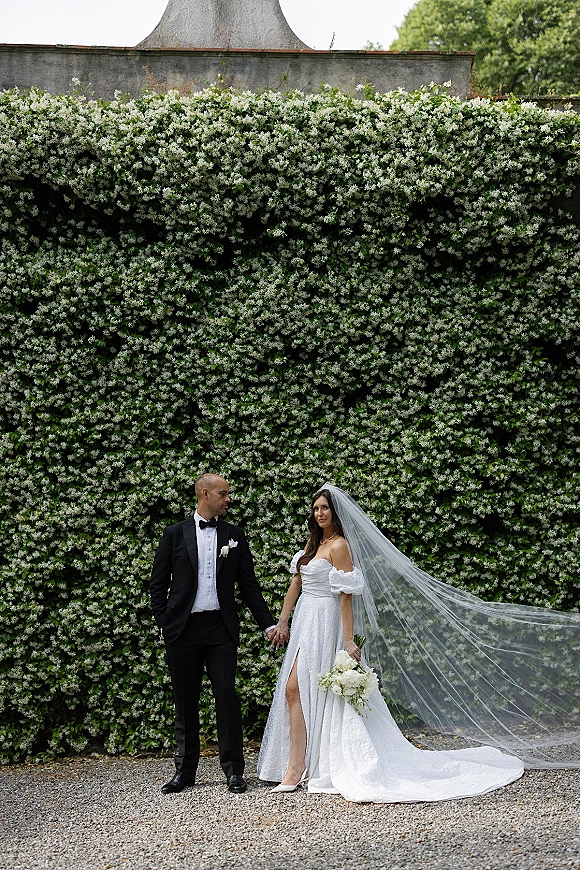 Couple portrait of bride and groom holding hands, her long veil blowing as they pose by a flowering ivy wall with bouquet