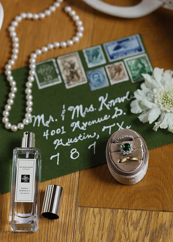 Wedding rings with emerald engagement ring in a ring box beside a calligraphy envelope, vintage stamps, pearls and perfume on a wood table