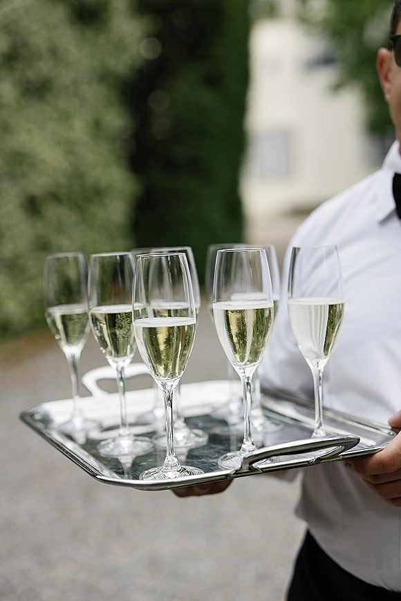 Champagne service with wedding welcome champagne flutes on a tray, held by a bow-tied server along a greenery-lined walkway outdoors