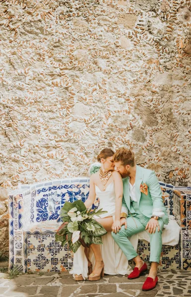 Couple portrait of bride and groom sitting close on a blue and white tile bench, bride holding bouquet against a textured stone wall