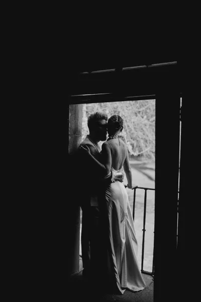 Wedding couple portrait, black and white wedding portrait of bride and groom embracing in a stone doorway with trees behind, satin open-back gown visible