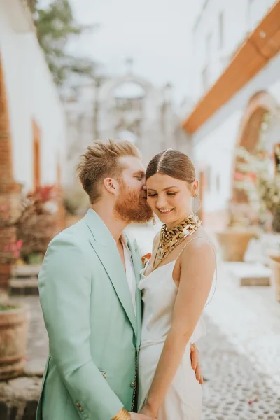 Couple portrait of groom kissing bride’s cheek, her satin wedding dress and hair accessory glowing in a white stucco courtyard with arches