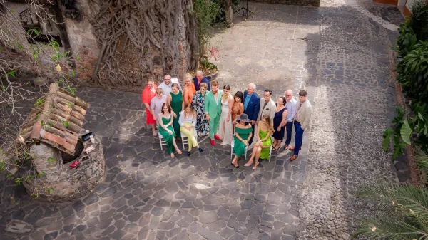 Wedding group photo of guests in cocktail attire, colorful dresses, and suits gathered in a stone courtyard by an ivy-covered wall