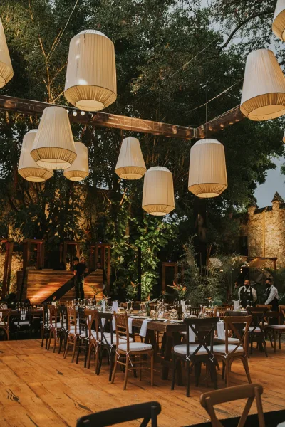 Outdoor reception decor with hanging lanterns and string lights over long wooden tables with white linens in a tree-lined courtyard