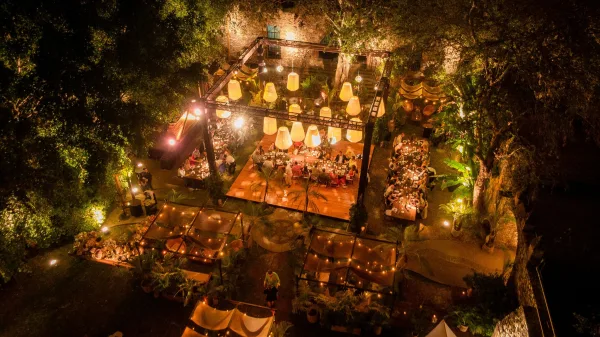 Outdoor wedding reception with string lights and hanging lanterns over banquet tables, candlelit lounge seating beneath trees by a stone wall at night