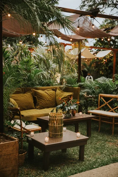 Outdoor lounge seating with a string light canopy over sofas, chairs, candles, and florals on a garden lawn under trees