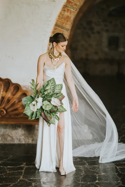 Bridal portrait of a modern bride in a simple satin wedding dress with long veil, holding a tropical leaf bouquet in an arched doorway