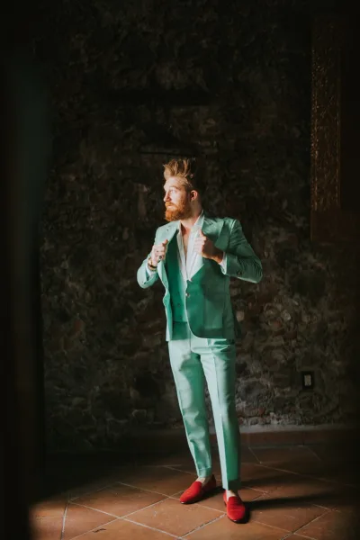Groom portrait in a mint green suit, adjusting his double-breasted jacket indoors by a stone wall and wooden door with red loafers