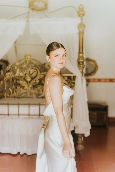 Bridal portrait in a satin slip wedding dress, bride looking over shoulder and holding her train beside a canopy bed with draped curtains