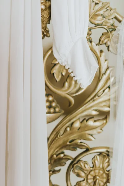 Wedding dress detail with a pearl button sleeve and tulle veil, shown close up against ornate gold wall decor and a white curtain