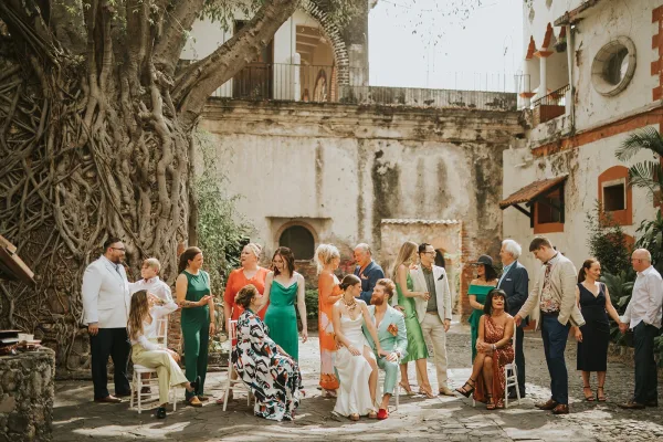 Wedding group portrait of the bride and groom with guests seated on white folding chairs in a stone courtyard, wide brim hat accent