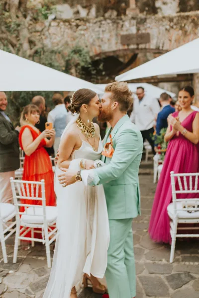 Wedding kiss as bride in veil and slip dress embraces groom in mint green suit, guests cheer in stone courtyard under shade sails