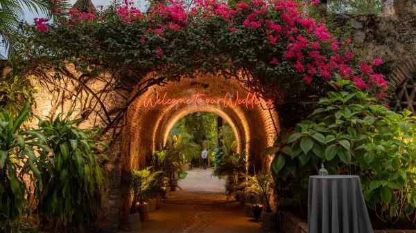 Wedding welcome sign with neon wedding sign glow, framed by bougainvillea and potted plants under a stone archway in a brick tunnel entrance