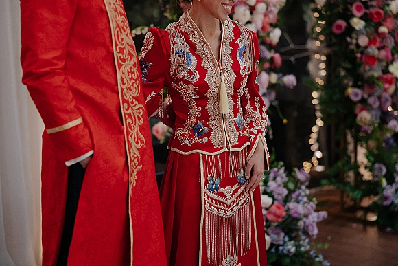 Wedding attire featuring a couple in red and gold embroidered bridal outfit and groom coat, posed before a floral backdrop with string lights