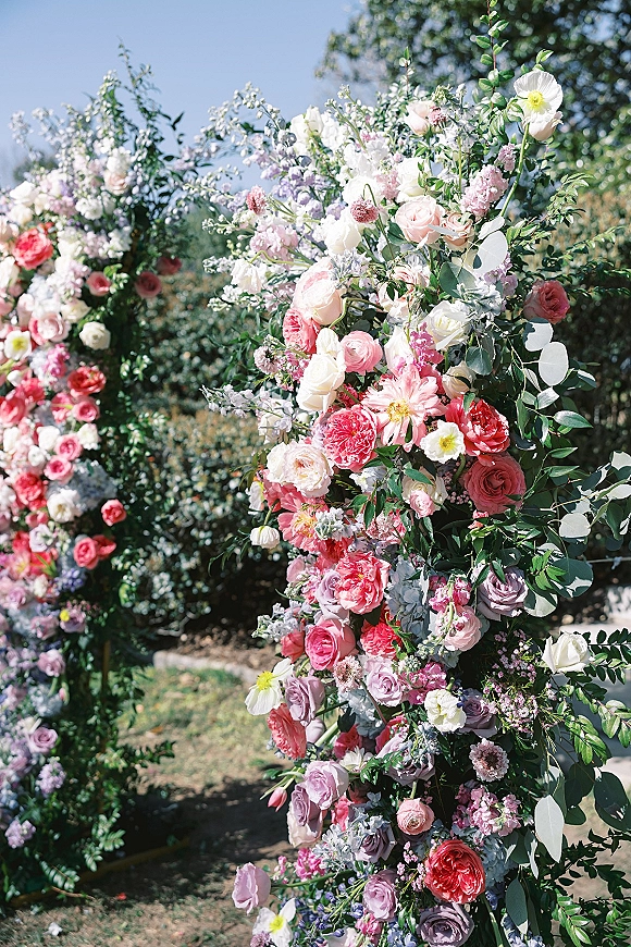 Floral wedding arch with pastel roses and ranunculus arranged asymmetrically, framed by greenery on a lawn beneath blue sky