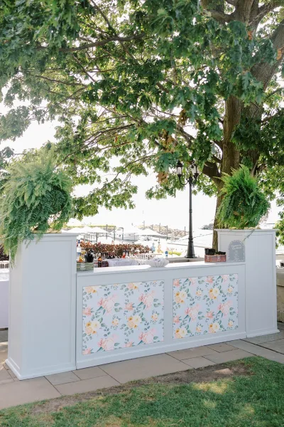 Wedding bar setup with a white outdoor wedding bar featuring floral panels, hanging ferns, and bottles on a stone patio by marina docks