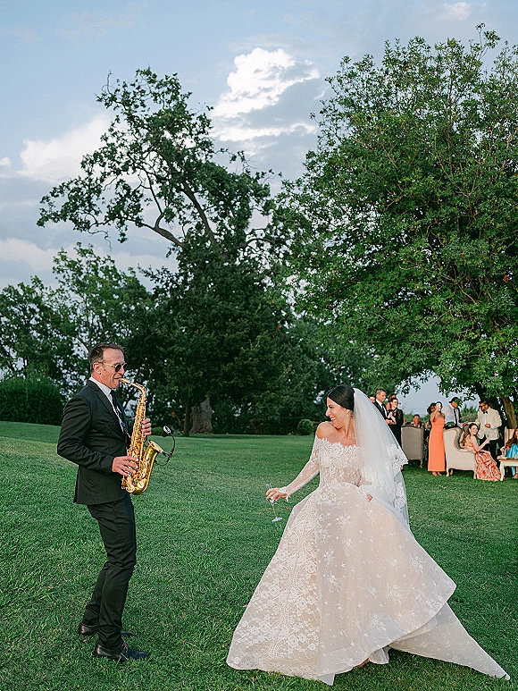 Wedding reception moment as a bride dances outdoors, laughing with a champagne flute beside a saxophonist in suit and sunglasses on a lawn