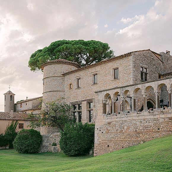 Stone wedding venue with stone arches and a tiled roof beside a green lawn and trees under a cloudy sky, tower rising above
