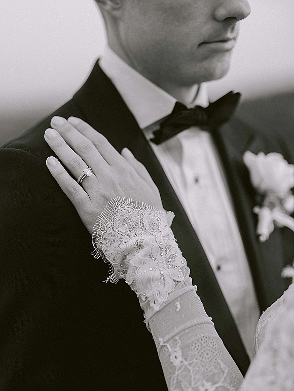 Wedding detail portrait of bride’s lace-gloved hand with engagement ring close-up resting on groom’s tuxedo lapel and boutonniere outdoors