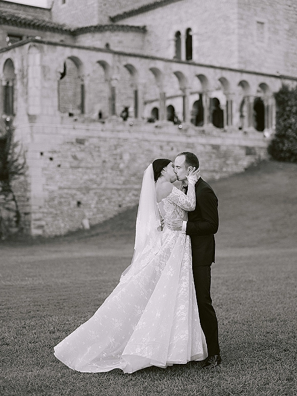 Wedding kiss portrait of bride and groom kissing, her long sleeve lace dress and cathedral veil flowing by stone arches on a lawn
