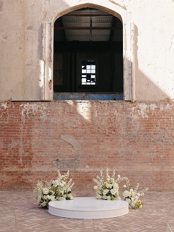 Wedding altar with round wedding stage on a white circular platform, framed by low white floral clusters against a brick wall arch