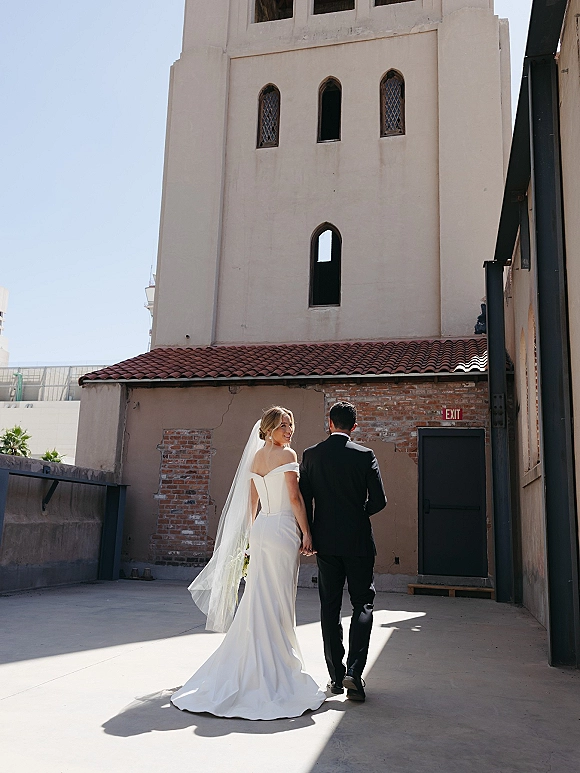 Couple portrait of newlyweds holding hands walking away as the bride looks back, her long veil trailing in a brick courtyard backdrop