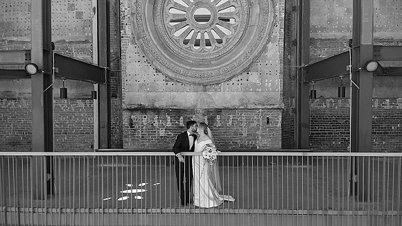 Wedding couple portrait of bride and groom kissing, bride holding bouquet with veil draped, leaning on railing by brick wall and circular window
