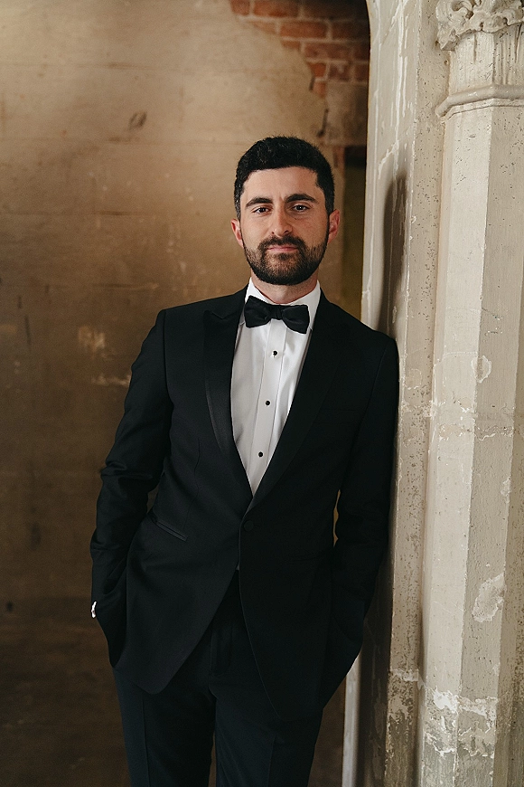 Groom portrait in a black tuxedo with bow tie, hands in pockets, standing by a stone archway and brick wall inside a historic building