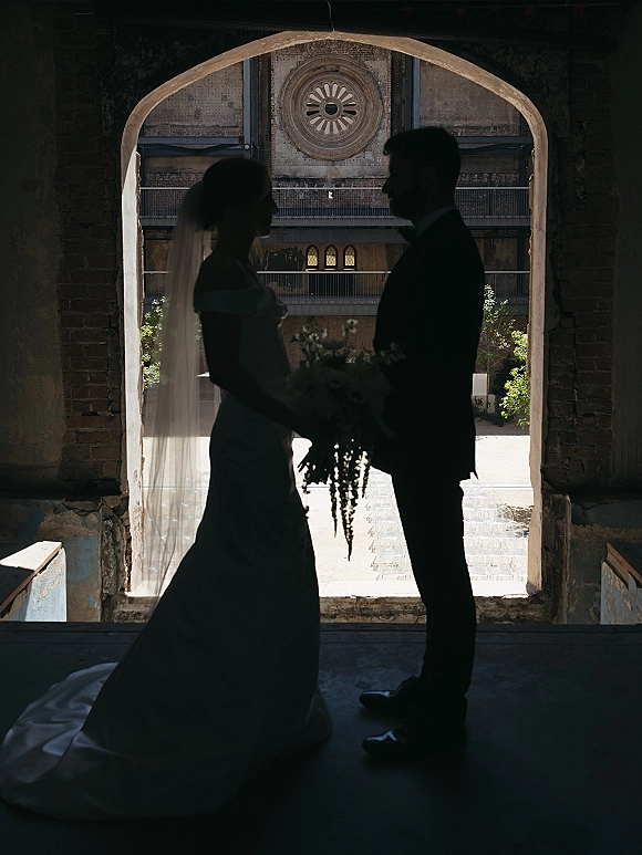 Couple portrait of bride and groom silhouette holding hands under a brick archway, backlit by sunlight with veil, bouquet, and tuxedo visible
