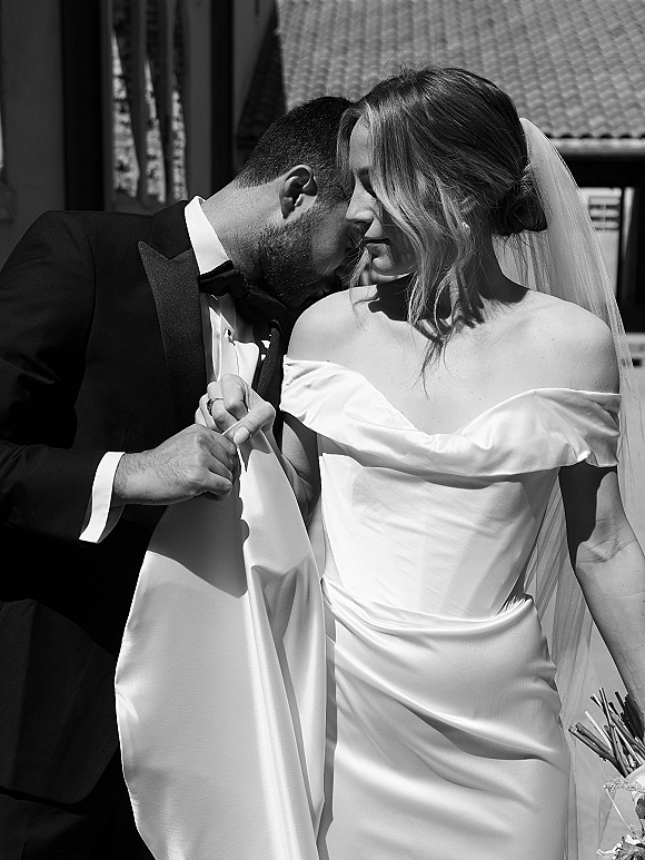 Couple portrait in a black and white wedding portrait, groom kissing bride’s forehead as they hold hands by a tile-roofed wall in sunlit shadows