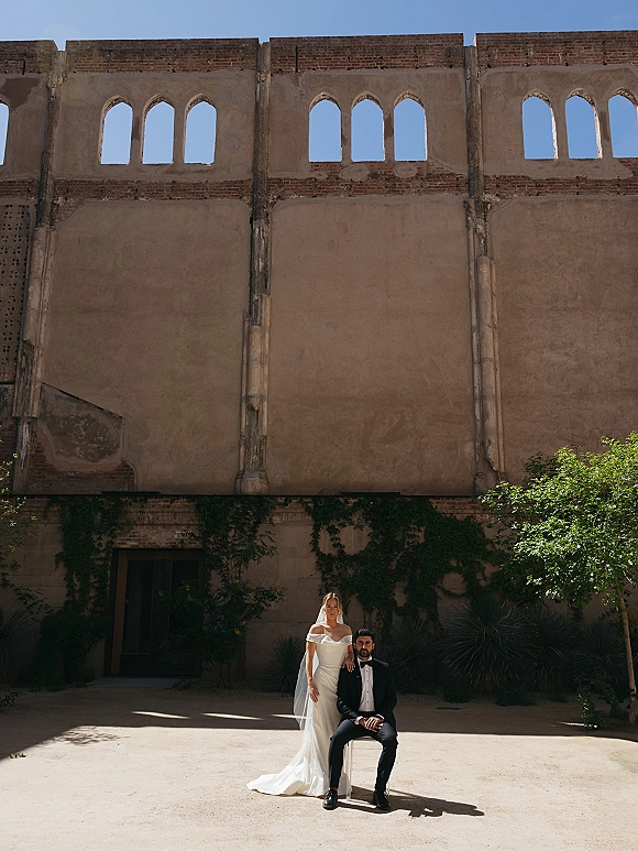Couple portrait of bride in off-the-shoulder wedding dress and veil beside groom in tuxedo on a chair in a stone courtyard with arched windows