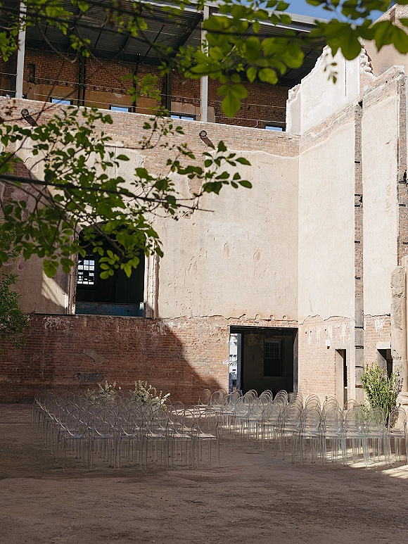 Ceremony setup with clear acrylic ceremony chairs in rows, simple aisle and greenery arrangements in an industrial brick courtyard with open roof