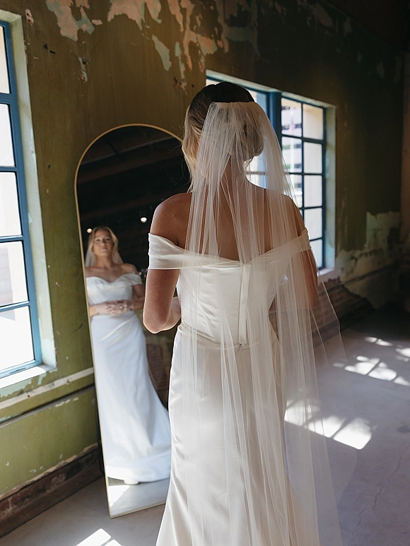 Bridal portrait of a bride looking in mirror, wearing an off shoulder wedding dress and long veil in an industrial room with arched doorway
