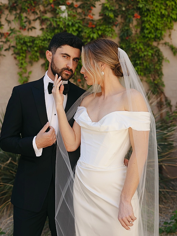 Couple portrait of bride in strapless wedding dress and veil touching groom’s face in black tuxedo, posed by an ivy stucco wall