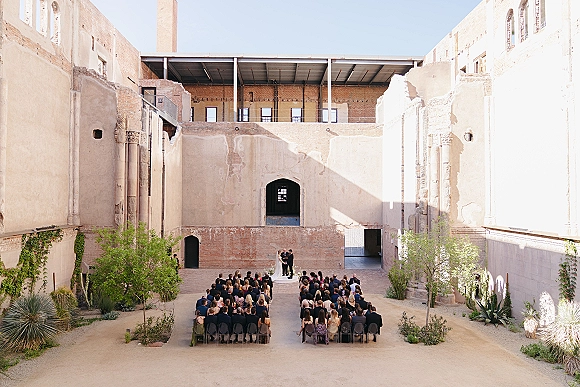 Wedding ceremony in an industrial courtyard with guests seated on chairs facing the bride and groom on a simple platform amid greenery shrubs