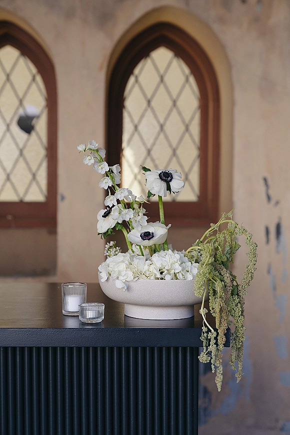 Wedding centerpiece with a modern floral centerpiece of white blooms and greenery in a low bowl, flanked by glass votives on a black pedestal table