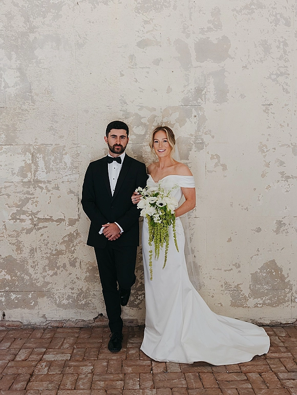 Couple portrait of bride and groom standing, bride in off-the-shoulder gown with veil holding a cascading bouquet before a textured plaster wall