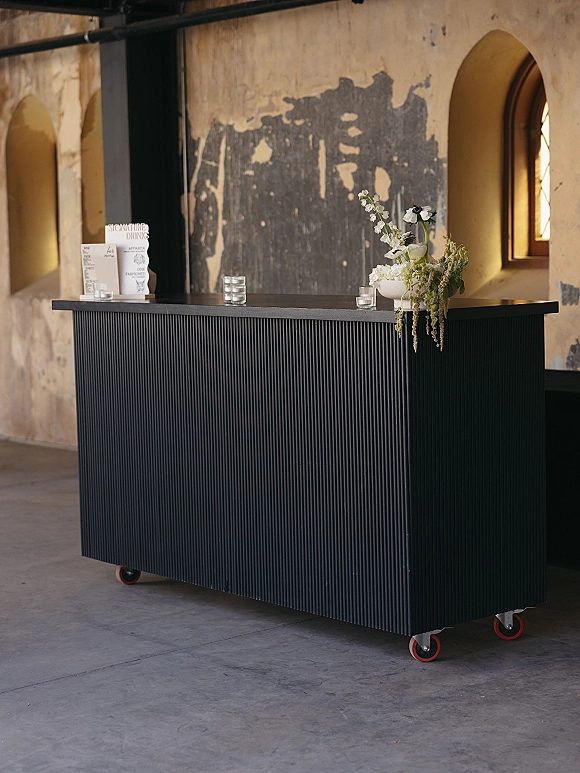 Wedding bar setup with a mobile wedding bar, drink menu sign, votive candles, and floral arrangement on a counter by an arched window niche