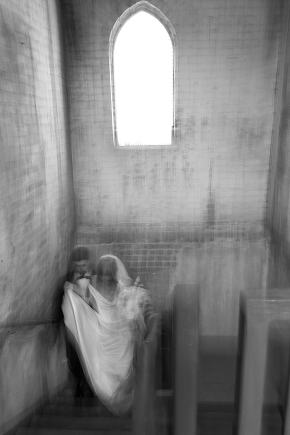 Couple portrait in a black and white wedding portrait style, bride in veil with bouquet beside groom in tuxedo in stone church pews