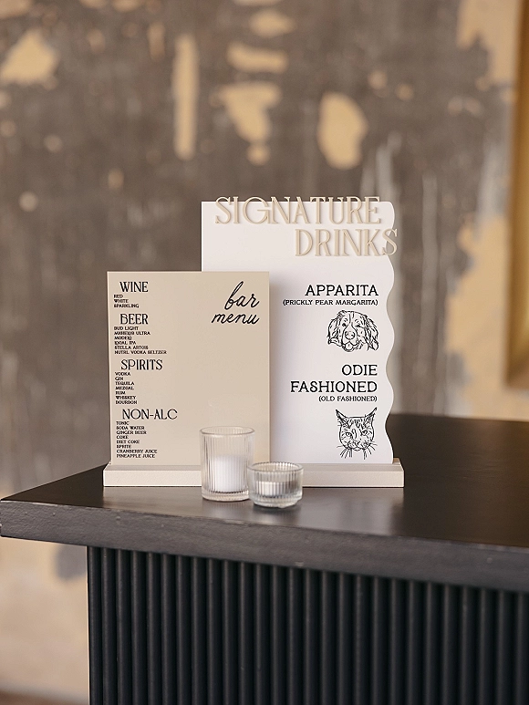 Wedding bar signage with a signature drinks sign in an acrylic holder on a bar counter, paired with votive candle holders against a textured wall