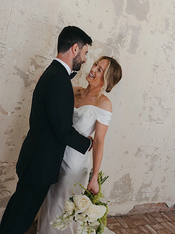 Couple portrait of bride and groom laughing as they embrace, her off-the-shoulder gown and white rose bouquet against a textured wall