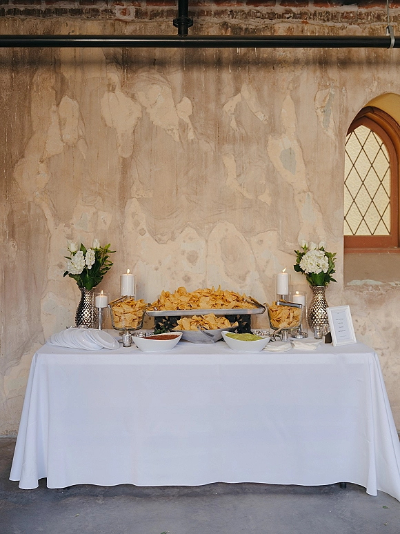 Wedding snack table with wedding chips and salsa bar, guacamole, tortilla chips, pillar candles, and white florals against a textured wall